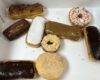 Assorted donuts and pastries in a white box, including a chocolate-glazed long doughnut, a white-iced pastry, a caramel-glazed éclair, a sprinkled donut, a plain sugar-coated doughnut, and a chocolate-glazed long doughnut on the side.