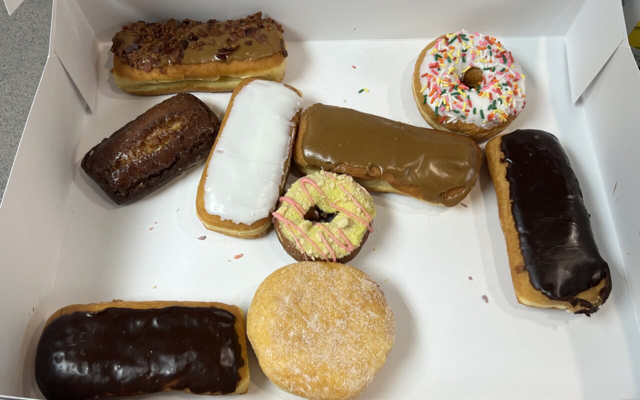 Assorted donuts and pastries in a white box, including a chocolate-glazed long doughnut, a white-iced pastry, a caramel-glazed éclair, a sprinkled donut, a plain sugar-coated doughnut, and a chocolate-glazed long doughnut on the side.