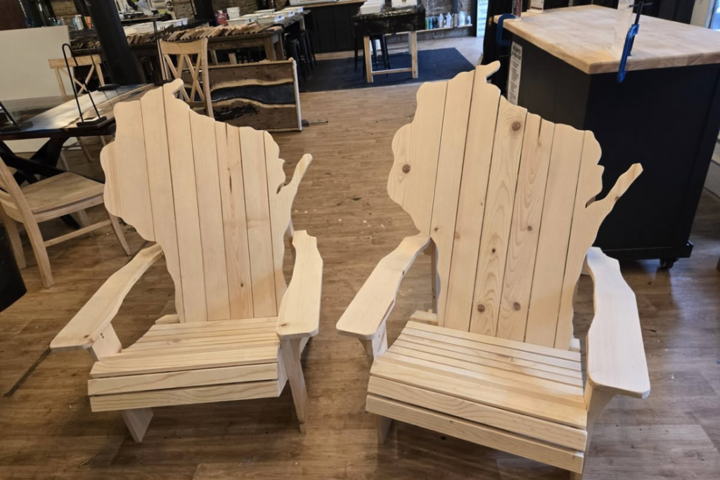 Two unfinished wooden Adirondack chairs shaped like the United States map, displayed in a workshop or store setting.