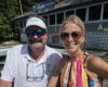Two smiling people posing at a dockside bar, man in a white polo with The Bub Water Trolley logo hat and glasses, woman in a colorful striped top and sunglasses.