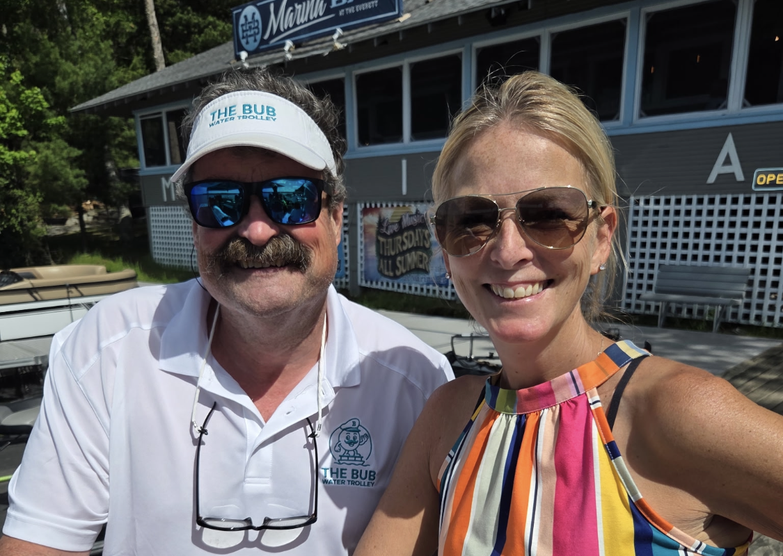 Two smiling people posing at a dockside bar, man in a white polo with The Bub Water Trolley logo hat and glasses, woman in a colorful striped top and sunglasses.