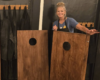 Smiling woman in a blue shirt stands between two wooden cornhole boards with circular holes indoors.