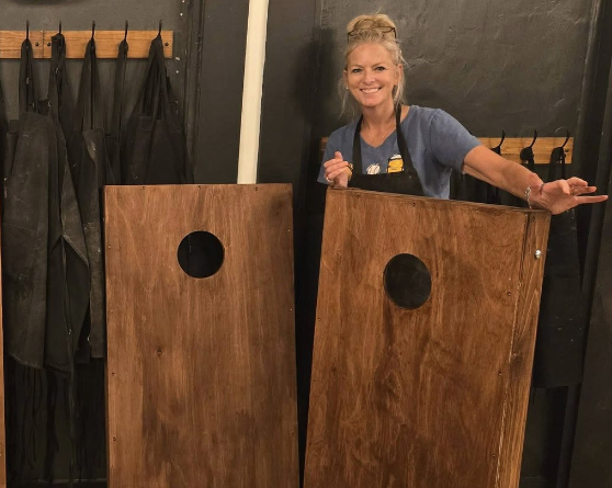 Smiling woman in a blue shirt stands between two wooden cornhole boards with circular holes indoors.