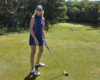 Woman golfer in a blue sleeveless dress and cap stands on a sunny golf course, holding a club and smiling at the camera.