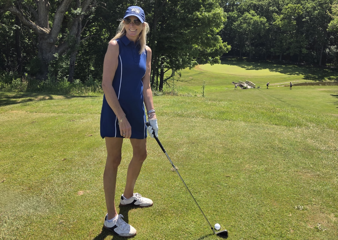 Woman golfer in a blue sleeveless dress and cap stands on a sunny golf course, holding a club and smiling at the camera.