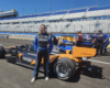Female race car driver in blue and black suit stands beside an orange IndyCar on a race track pit lane, grandstands in background.