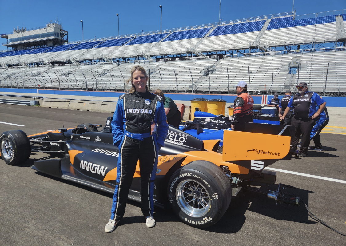 Female race car driver in blue and black suit stands beside an orange IndyCar on a race track pit lane, grandstands in background.