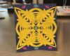 Yellow paper-cut mandala on a navy card, with a green center star, orange dots, and pink/teal corner accents.