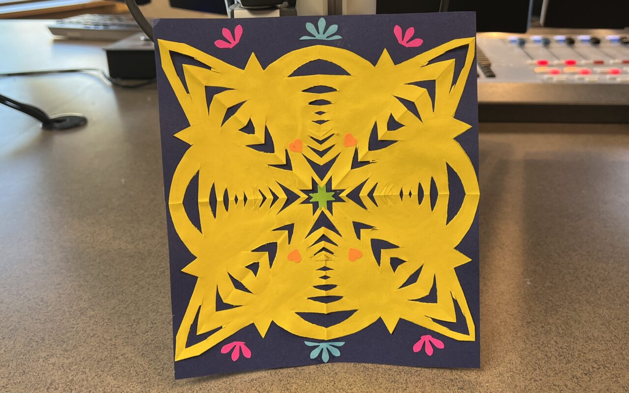 Yellow paper-cut mandala on a navy card, with a green center star, orange dots, and pink/teal corner accents.