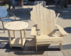 Outdoors: unfinished wooden Adirondack chair beside a round wooden side table on a paved patio; a small white dog in the background.