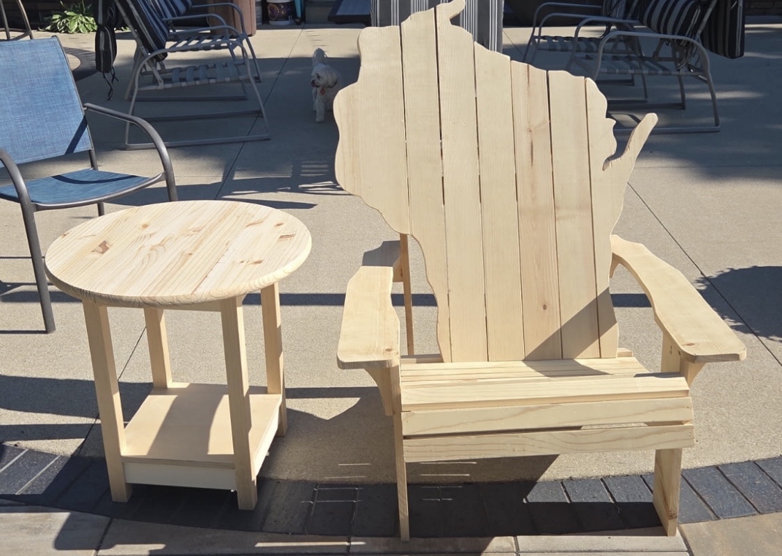 Outdoors: unfinished wooden Adirondack chair beside a round wooden side table on a paved patio; a small white dog in the background.