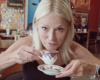 Woman with blonde hair at a cafe, about to sip from an ornate teacup held near her lips, saucer in the other hand.