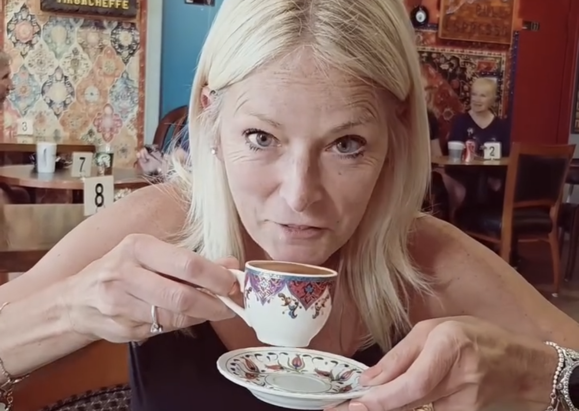 Woman with blonde hair at a cafe, about to sip from an ornate teacup held near her lips, saucer in the other hand.