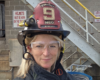 Female firefighter wearing a helmet marked with the number 9, clear safety goggles, and turnout gear outdoors by a metal staircase.