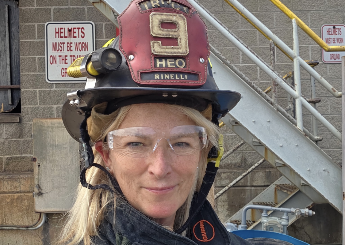 Female firefighter wearing a helmet marked with the number 9, clear safety goggles, and turnout gear outdoors by a metal staircase.