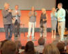 Six people on a stage, speaking into microphones in front of an audience.
