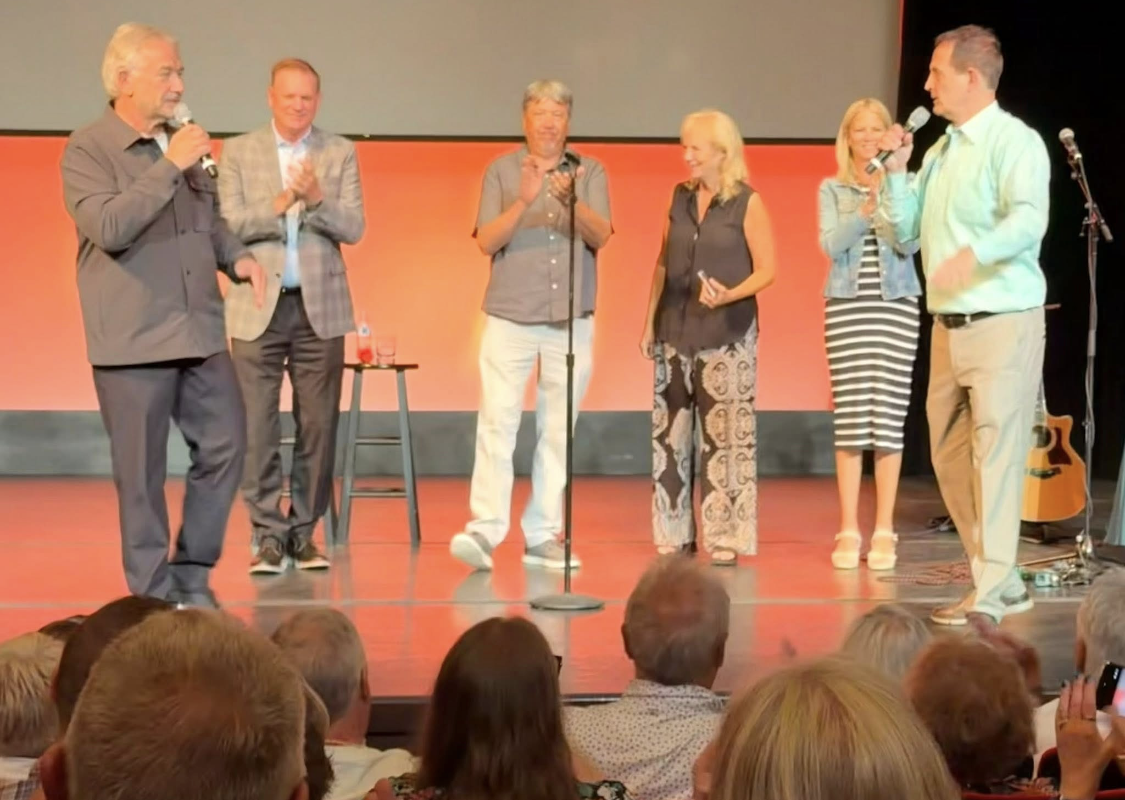 Six people on a stage, speaking into microphones in front of an audience.
