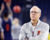 Older male basketball coach wearing a gray jacket with an orange 'S' logo, arms crossed, serious expression on a court.