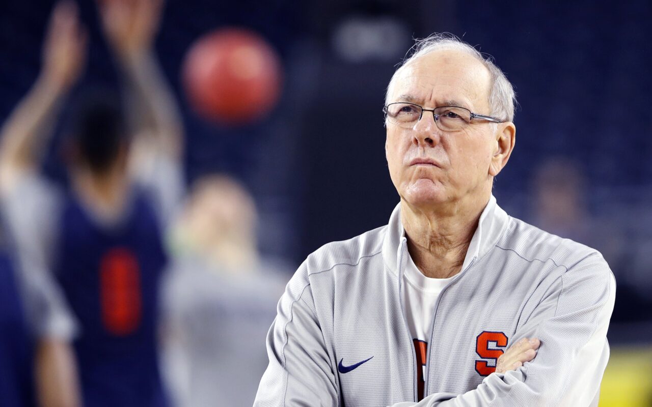 Older male basketball coach wearing a gray jacket with an orange 'S' logo, arms crossed, serious expression on a court.
