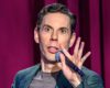 Male comedian on stage with microphone, wide-eyed expression and raised hand near mouth against maroon curtains
