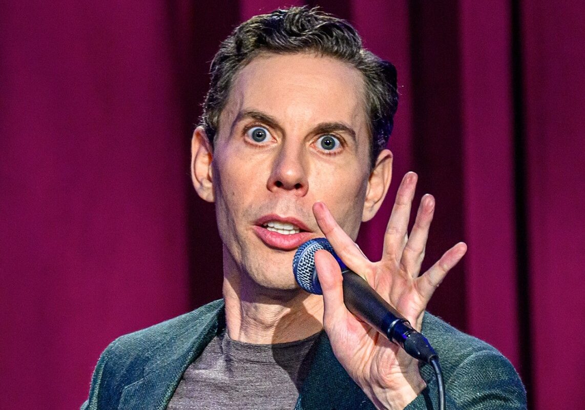 Male comedian on stage with microphone, wide-eyed expression and raised hand near mouth against maroon curtains