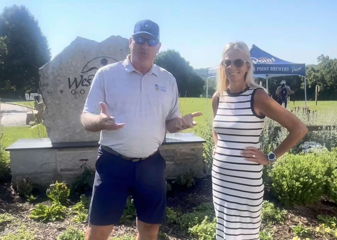 Two adults posing outdoors by a stone sign at a park event, with a blue canopy on the right and greenery around.