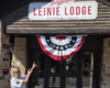 Woman in sunglasses points at the Leinie Lodge sign above a doorway with patriotic bunting.