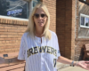 Woman with blonde hair and sunglasses wearing a Brewers jersey, standing outside a brick building with arms outstretched.