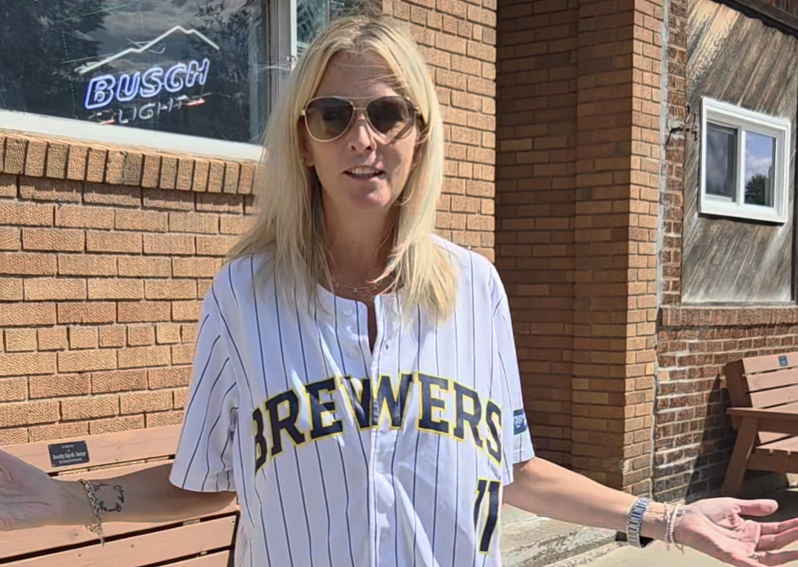 Woman with blonde hair and sunglasses wearing a Brewers jersey, standing outside a brick building with arms outstretched.