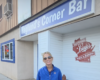 Woman in a blue jacket and sunglasses stands outside Haglund's Corner Bar, smiling near a Wisconsin Levi's mural on the wall.