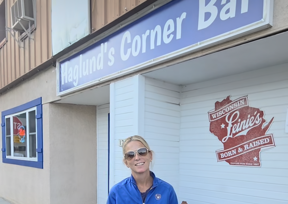 Woman in a blue jacket and sunglasses stands outside Haglund's Corner Bar, smiling near a Wisconsin Levi's mural on the wall.