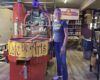 Woman in a blue t-shirt stands beside a large red coffee roasting machine in a roastery, with a Café De Arts banner nearby.