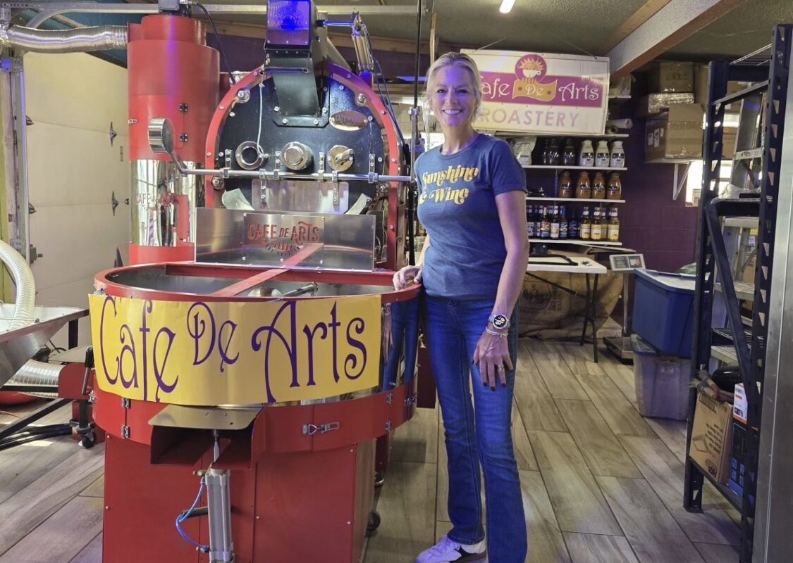Woman in a blue t-shirt stands beside a large red coffee roasting machine in a roastery, with a Café De Arts banner nearby.