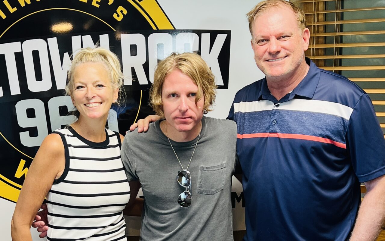 Three adults posing together in a radio studio, left to right: a woman in a black-and-white striped sleeveless top, a man in a gray t-shirt with sunglasses hanging from his shirt, and a man in a blue polo with white and red stripes, standing in front of a large circular station sign.