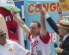 Victory celebration: man in a red-and-white jersey raises both arms while two others cheer, a hot-dog mascot on the left and a host with a microphone on the right, colorful banners in the background.