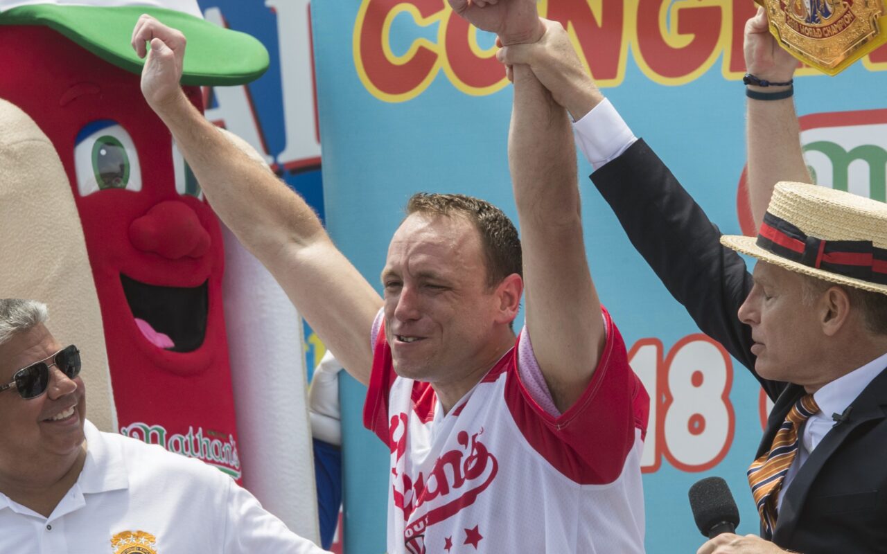 Victory celebration: man in a red-and-white jersey raises both arms while two others cheer, a hot-dog mascot on the left and a host with a microphone on the right, colorful banners in the background.