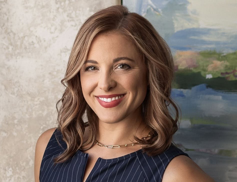 Smiling woman with shoulder-length light brown hair wearing a navy pinstripe sleeveless top and a gold chain necklace.