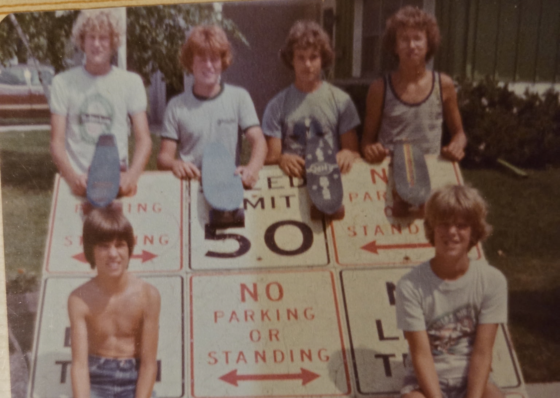 Six teenagers posing with skateboards on top of large 'No Parking or Standing' signs outdoors.