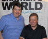Two men pose for a photo indoors in front of a backdrop that spells 'WORLD'; the man on the left wears a blue striped polo and the man on the right sits in a black shirt.
