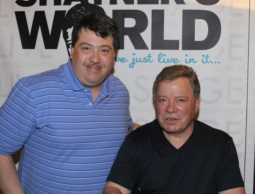 Two men pose for a photo indoors in front of a backdrop that spells 'WORLD'; the man on the left wears a blue striped polo and the man on the right sits in a black shirt.