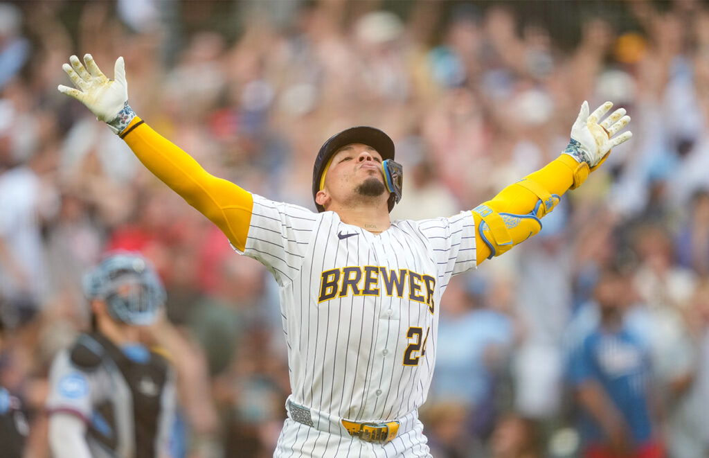 Baseball player in Brewers uniform raises arms in celebration as fans cheer in the background.